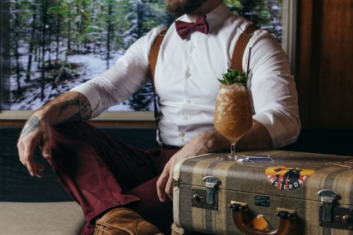 A man in a bow tie and suspenders sits beside a vintage suitcase, with a cocktail garnished with mint in hand.