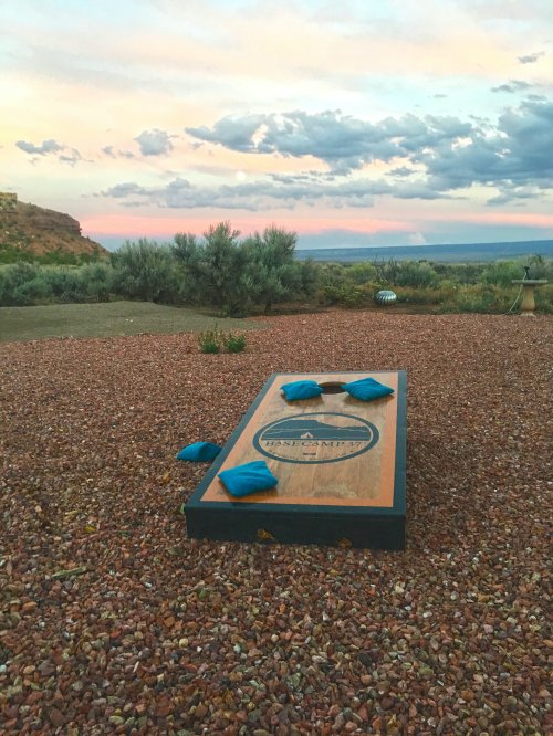 A cornhole game set on gravel with blue bags, surrounded by shrubs and a colorful sunset sky.