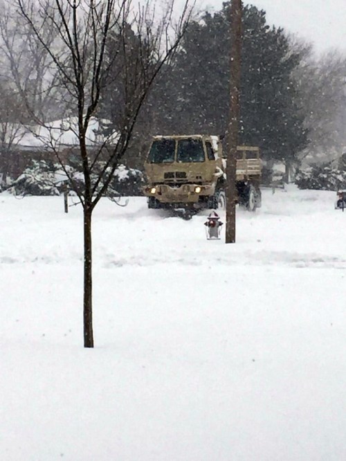 A military vehicle parked in a snowy landscape, with trees and a lamppost visible in the foreground.