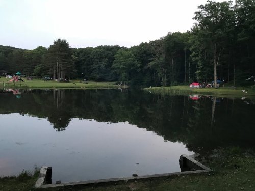 A serene lake surrounded by trees, reflecting the evening sky, with tents and playground equipment in the background.