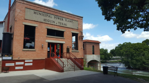 Historic Municipal Power Plant building in Seguin, Texas, with a river and trees in the background under a blue sky.