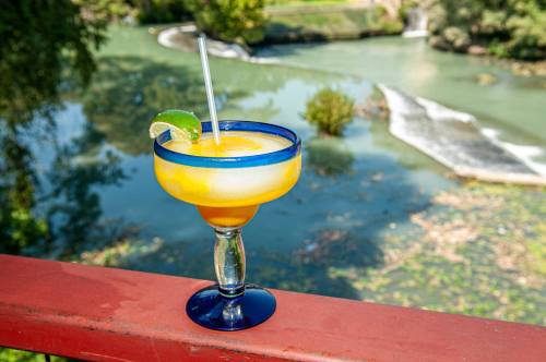 A vibrant margarita with a lime wedge sits on a railing, overlooking a serene river landscape.