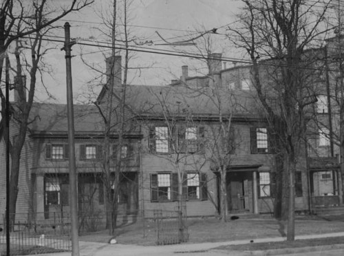 Black and white photo of a two-story house with trees and power lines in front, showcasing a vintage architectural style.