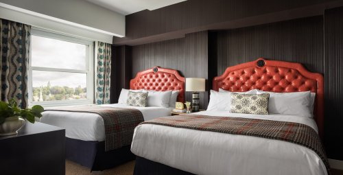 A stylish hotel room featuring two beds with tufted red headboards, patterned bedding, and a large window with a view.