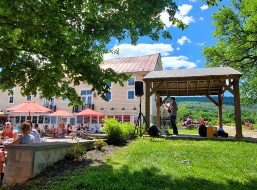 A performer sings on a stage in a sunny outdoor setting, with people dining and enjoying the view of green hills.