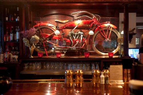 A vintage red Indian motorcycle displayed on a wooden bar shelf, surrounded by decorative flowers and glassware.