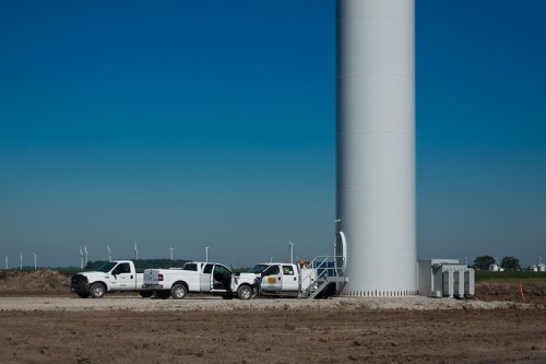 Two white trucks parked near a large wind turbine tower on a clear day, with wind turbines visible in the background.