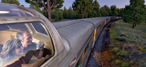 A couple enjoys a scenic train ride, looking out from the front window as the train travels through a forested area.