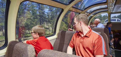 Two boys sit in a train car, looking out the window at a scenic view, while adults chat in the background.