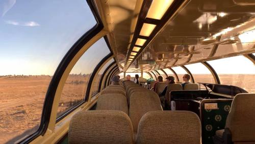 Interior of a train with large windows, showing passengers seated and a view of a vast, open landscape outside.
