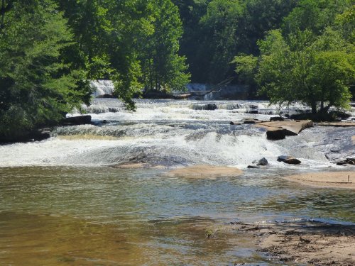 A serene river with cascading waterfalls surrounded by lush green trees and rocky banks under a clear blue sky.