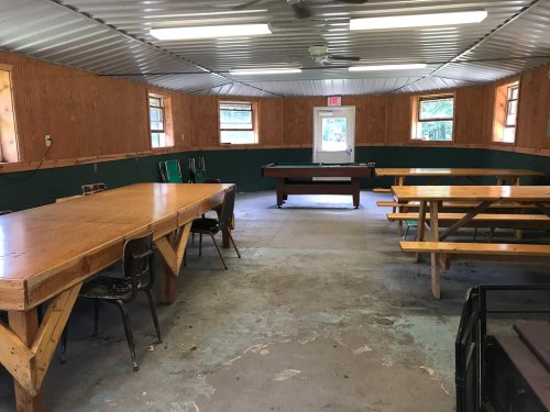 Interior of a rustic room with wooden tables, chairs, and a pool table, featuring green walls and large windows.