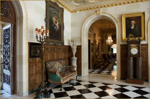 Elegant hallway with checkered floor, ornate furniture, and classic artwork, featuring chandeliers and decorative wood paneling.
