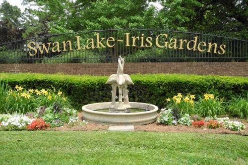 Sign for Swan Lake-Iris Gardens with a fountain and colorful flowers in the foreground. Lush greenery surrounds the area.