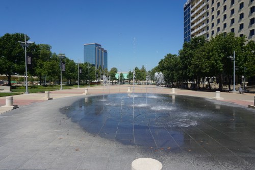 A sunny plaza with a fountain, surrounded by trees and modern buildings in the background.