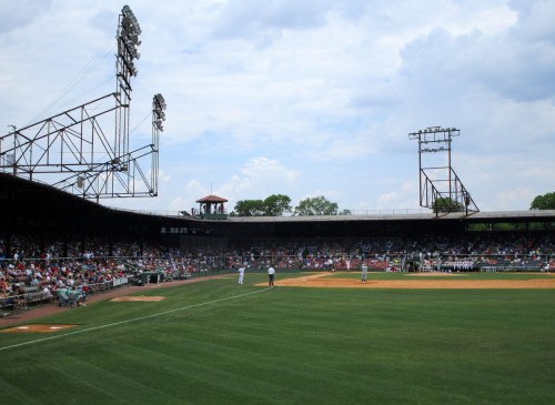A baseball field filled with spectators, featuring players on the field and a cloudy sky above.