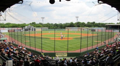 A wide view of a baseball field with players on the diamond and spectators in the stands under a cloudy sky.