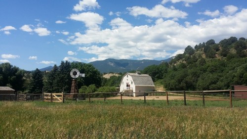 A scenic farm with a white barn, windmill, and lush green fields, set against a backdrop of mountains and blue sky.