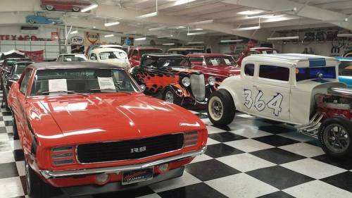 A vintage car museum featuring classic cars, including a red Camaro and various hot rods on a checkered floor.