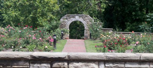 A stone archway leads into a vibrant rose garden, surrounded by lush greenery and a brick pathway.