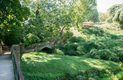 A serene park scene featuring a stone bridge over a lush, green area with trees and steps in the background.