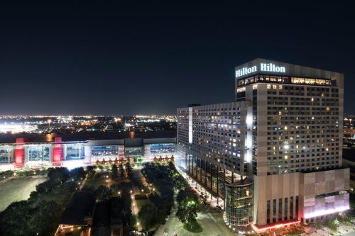 Aerial view of the Hilton hotel at night, illuminated against a city skyline with lights in the background.