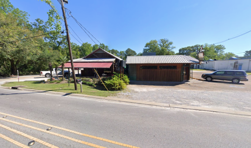 A small roadside building with a metal roof, surrounded by trees and parked cars on a sunny day.