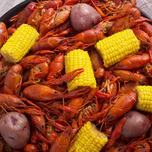 A close-up of a seafood boil featuring red crawfish, yellow corn, and purple potatoes on a black platter.