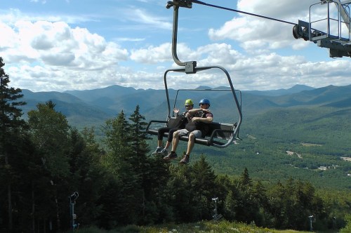 Two people on a ski lift, surrounded by lush green mountains and a blue sky with fluffy clouds.