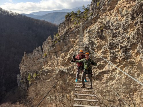 Two climbers on a narrow suspension bridge over a rocky gorge, surrounded by mountains and trees.