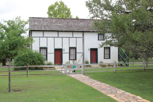 A two-story historic house with a stone path, green lawn, and trees, featuring red doors and a wooden fence.