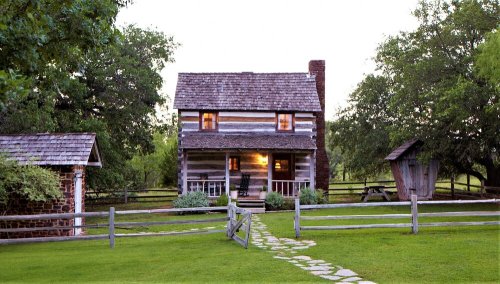 A rustic log cabin with a stone chimney, surrounded by greenery and a wooden fence, at dusk.