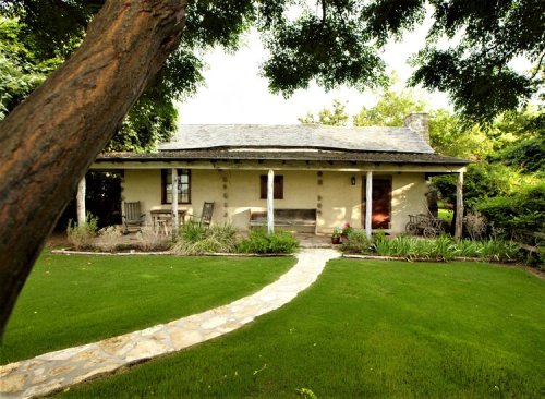 A charming cottage with a stone pathway, surrounded by lush greenery and trees, featuring a porch and outdoor seating.