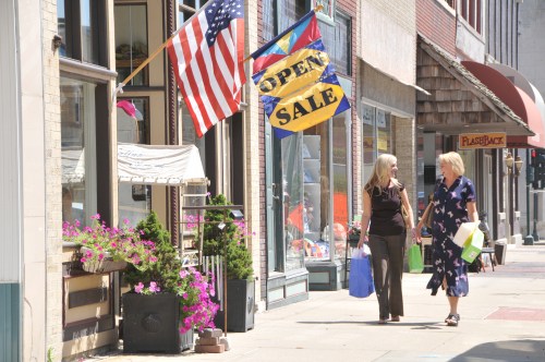 Two women walk along a sunny street, carrying shopping bags, with flags and flowers in the background.
