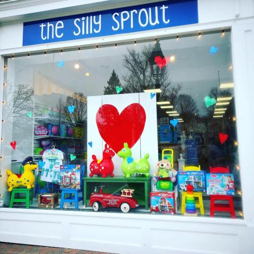 Colorful storefront display featuring toys, including green and red animals, with hearts and a large red heart backdrop.