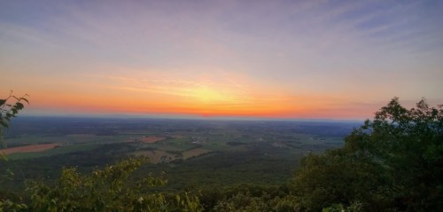 A vibrant sunset over rolling hills and fields, with a clear sky transitioning from orange to blue.