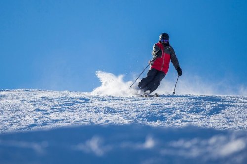 A skier in a red jacket glides down a snowy slope against a clear blue sky, creating a spray of snow.
