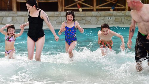 A family enjoys splashing in a pool, with two children and two adults playing in the water.