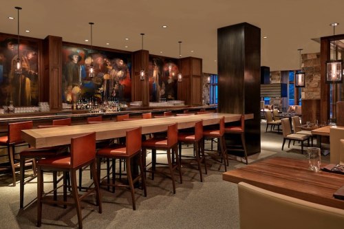 Modern bar interior with wooden tables, high chairs, and artistic wall decor, creating a warm, inviting atmosphere.