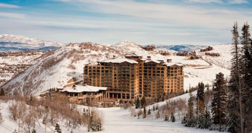 A large, modern hotel nestled in snowy mountains, surrounded by trees and a clear blue sky.