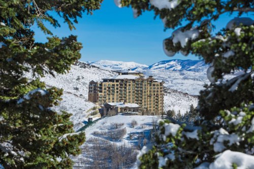 A snowy mountain landscape featuring a large hotel nestled among trees, with blue skies and distant peaks.