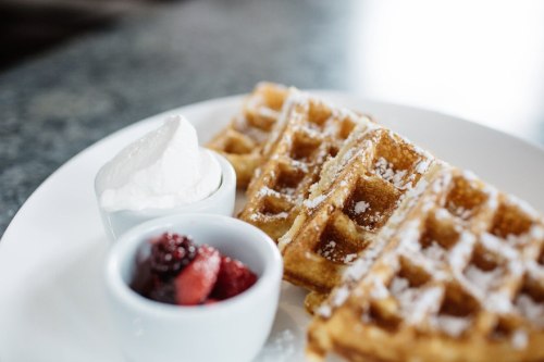 A plate of golden waffles topped with powdered sugar, served with whipped cream and a side of mixed berries.