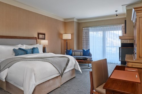 A cozy hotel room featuring a king bed, desk, seating area, and large windows with natural light.