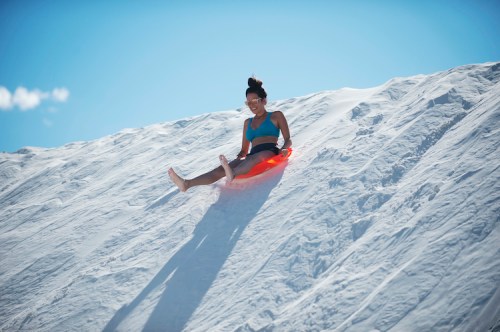 A person in a blue top sleds down a snowy hill on a bright orange sled under a clear blue sky.