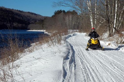 A person rides a yellow snowmobile along a snowy path by a river, surrounded by trees and mountains.