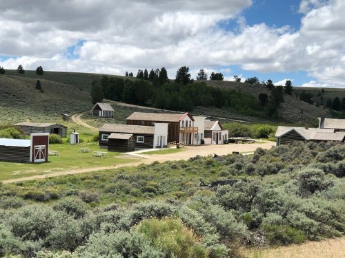 A rustic western-style town with wooden buildings, surrounded by green hills and cloudy skies.