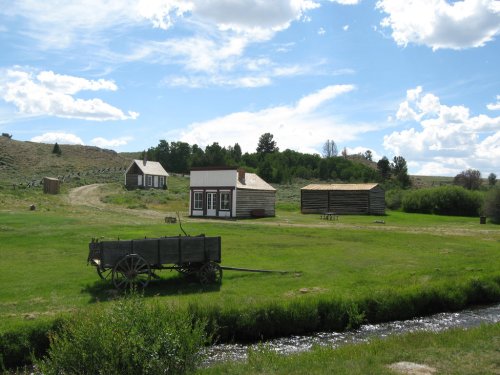 A historic settlement with wooden buildings, a wagon, and a stream, set against a blue sky with fluffy clouds.