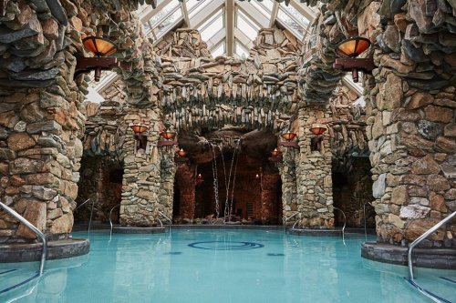 A luxurious indoor pool surrounded by stone walls and decorative lighting, featuring a waterfall and glass ceiling.