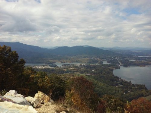 A scenic view of mountains and a lake under a cloudy sky, with trees in the foreground and a winding road in the distance.
