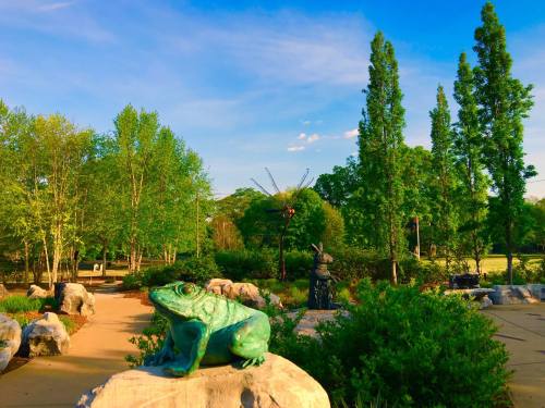 A green frog sculpture among rocks in a lush park with trees and a blue sky in the background.